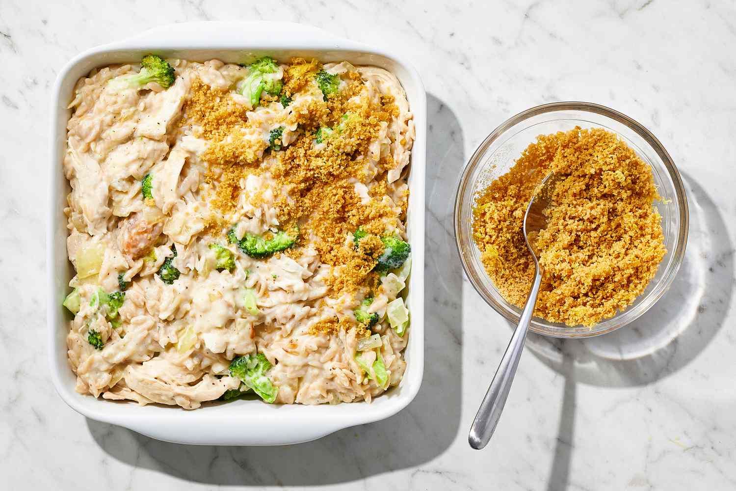 overhead view of casserole in a dish with breadcrumb mixture sprinkled on half of the top and a bowl of breadcrumb mixture with a spoon in it