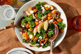 Serving bowl of autumn salad with utensils on a rust colored linen, surrounded by glasses of sparkling water. 
