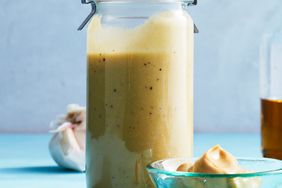 Jar of Lemon-Garlic Vinaigrette with mustard in a bowl in the foreground and garlic cloves. 