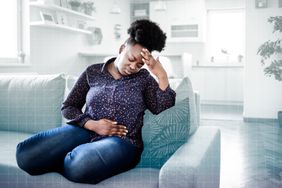 a photo of a woman sitting on her couch with a hand up to her head and her other hand holding her stomach