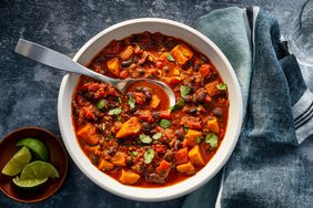 Sweet potato & black bean chili in a white bowl with a spoon in it. Lime wedges on the side and fresh cilantro sprinkled on top