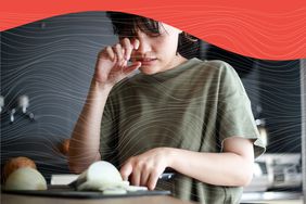 Woman rubbing her eye while cutting an onion on a kitchen counter against a gray background